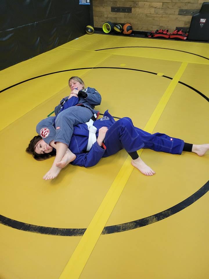 One training partner in a grey gi controls another in a blue gi on the yellow mats