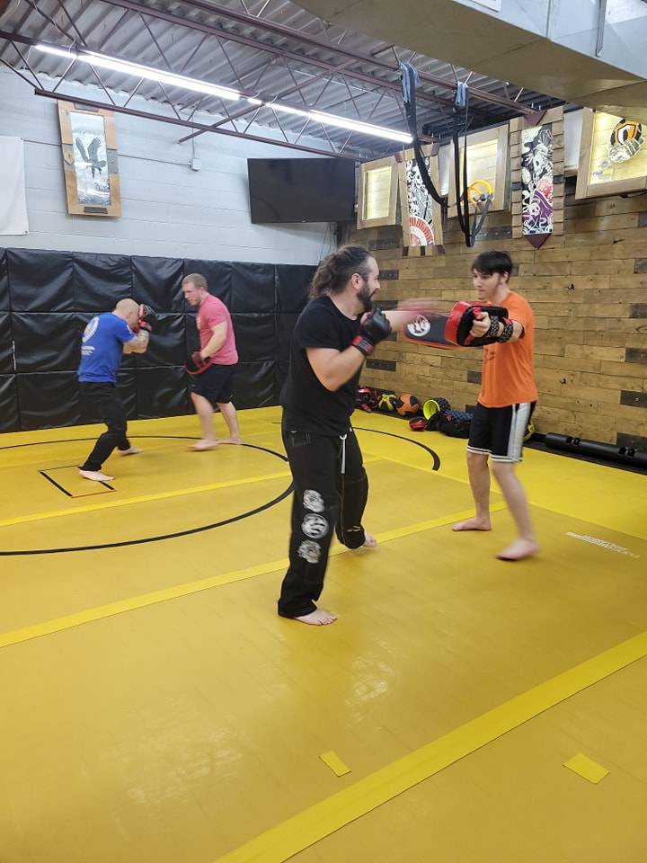 A student in an orange shirt holds pads while a boxer in black throws a punch, with two more pairs training behind them