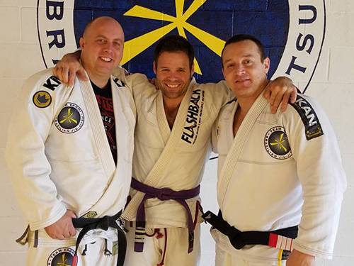 Three black belt coaches in gis with their arms around each other, in front of the Team Vaghi logo