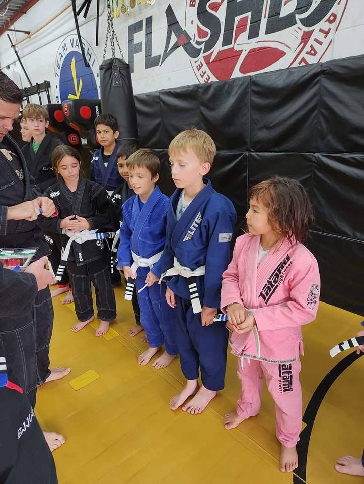 A row of kids in blue, black and pink gis waits barefoot on the mat while a coach hands out new belts