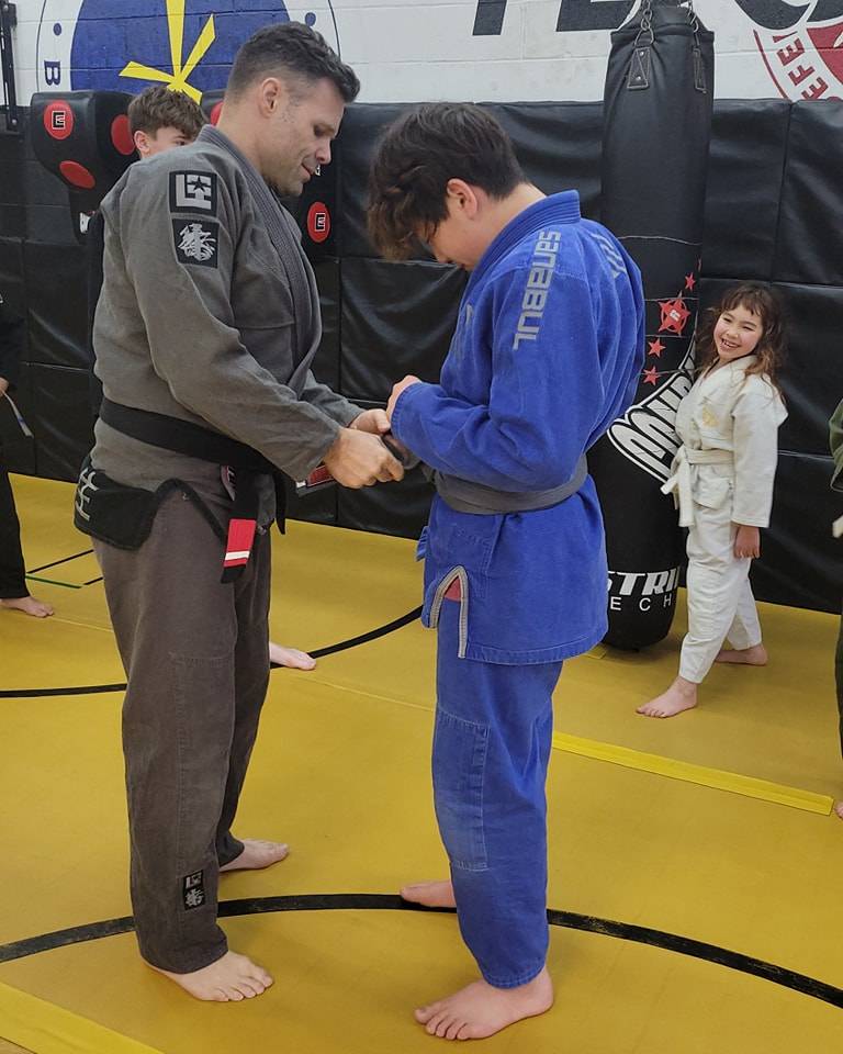 A coach in a grey gi ties a new belt on a young student in a blue gi while a smiling girl in a white gi watches
