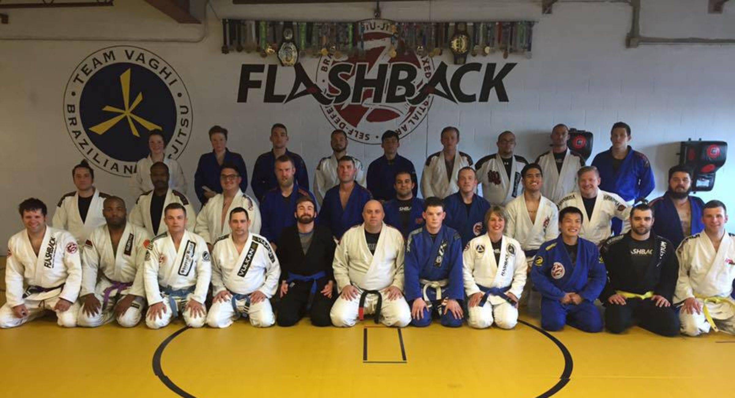 About forty Flashback members in white, blue and black gis lined up on the yellow mats, under the Flashback and Team Vaghi logos on the wall